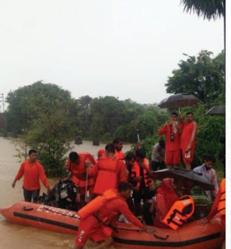 An NDRF rescue crew in orange MM life jackets on an inflatable boat in floodwater